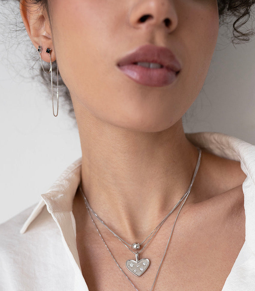 Close-up of a woman wearing a silver necklace with heart and teardrop pendants, against a neutral background.