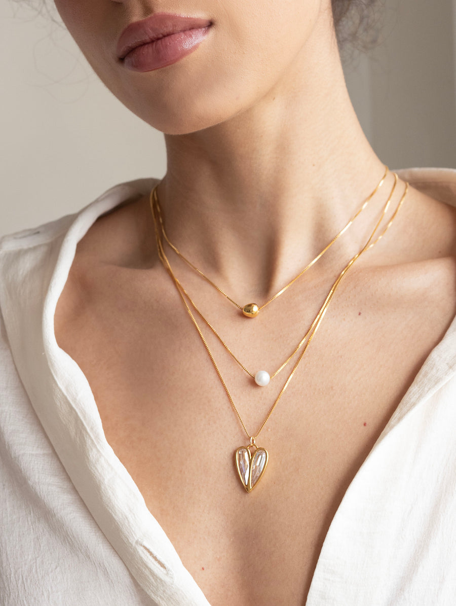 Close-up of a person wearing gold necklaces with heart and pearl pendants on a neutral background